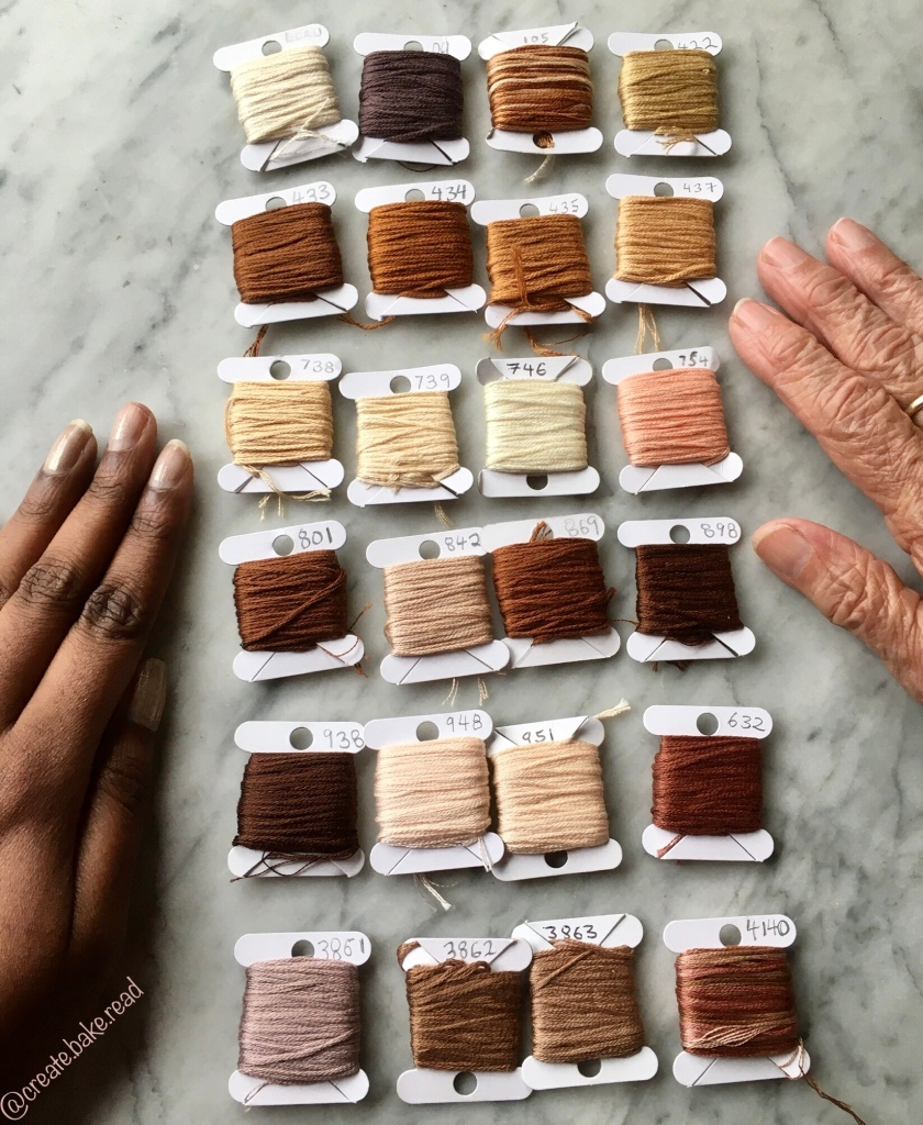 A brown person’s hand  on the left side and a white person’s hand on the right side. With the following DMC floss threads on cardboard bobbins in the middle:
Ecru, 09, 105, 422,
433, 434, 435, 437,
632, 738, 739, 746,
754, 801, 842, 869,
898,938, 948, 951,
3861, 3862, 3863, 4140.
Light brown watermark reads: @create.bake.read
On a grey marble table.